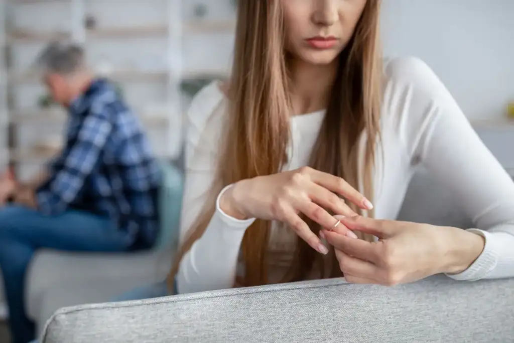 Frau dreht nachdenklich ihren Ehering, der Partner sitzt abgewandt im Hintergrund – eine Trennung kostet mehr als eine Paartherapie